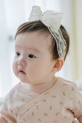 Baby girl wearing Avauma Rumiere Ribbon Band and pink floral wrap bodysuit, looking to the side indoors